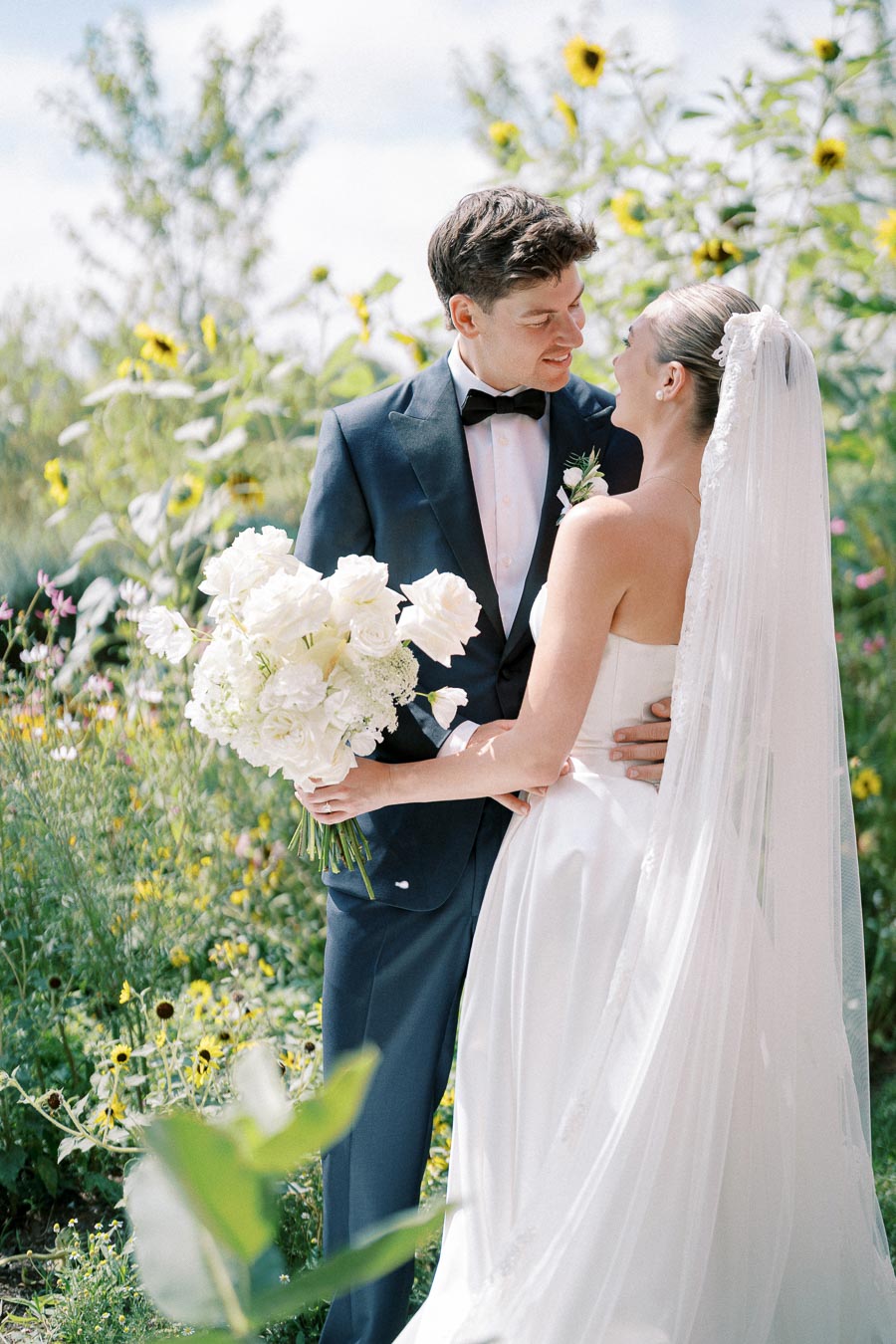 Elegant bride and groom embracing in a vibrant garden setting with wildflowers, bride holding a bouquet of white flowers, wearing a long veil and white gown, groom in a navy suit and bow tie, celebrating their wedding day outdoors.