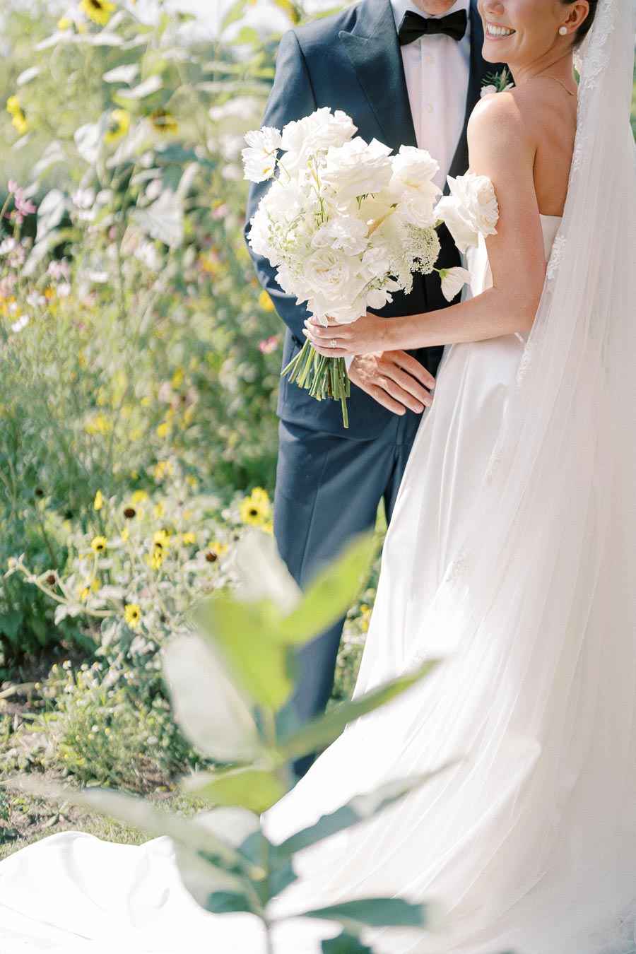 A bride in a white wedding dress holds a bouquet of white flowers, standing next to a groom in a dark suit, set in a picturesque garden filled with greenery and flowers.