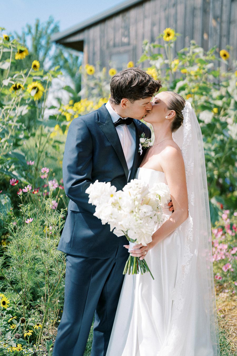 Bride and groom kissing in a garden with sunflowers, bride holding white bouquet, outdoor wedding scene.
