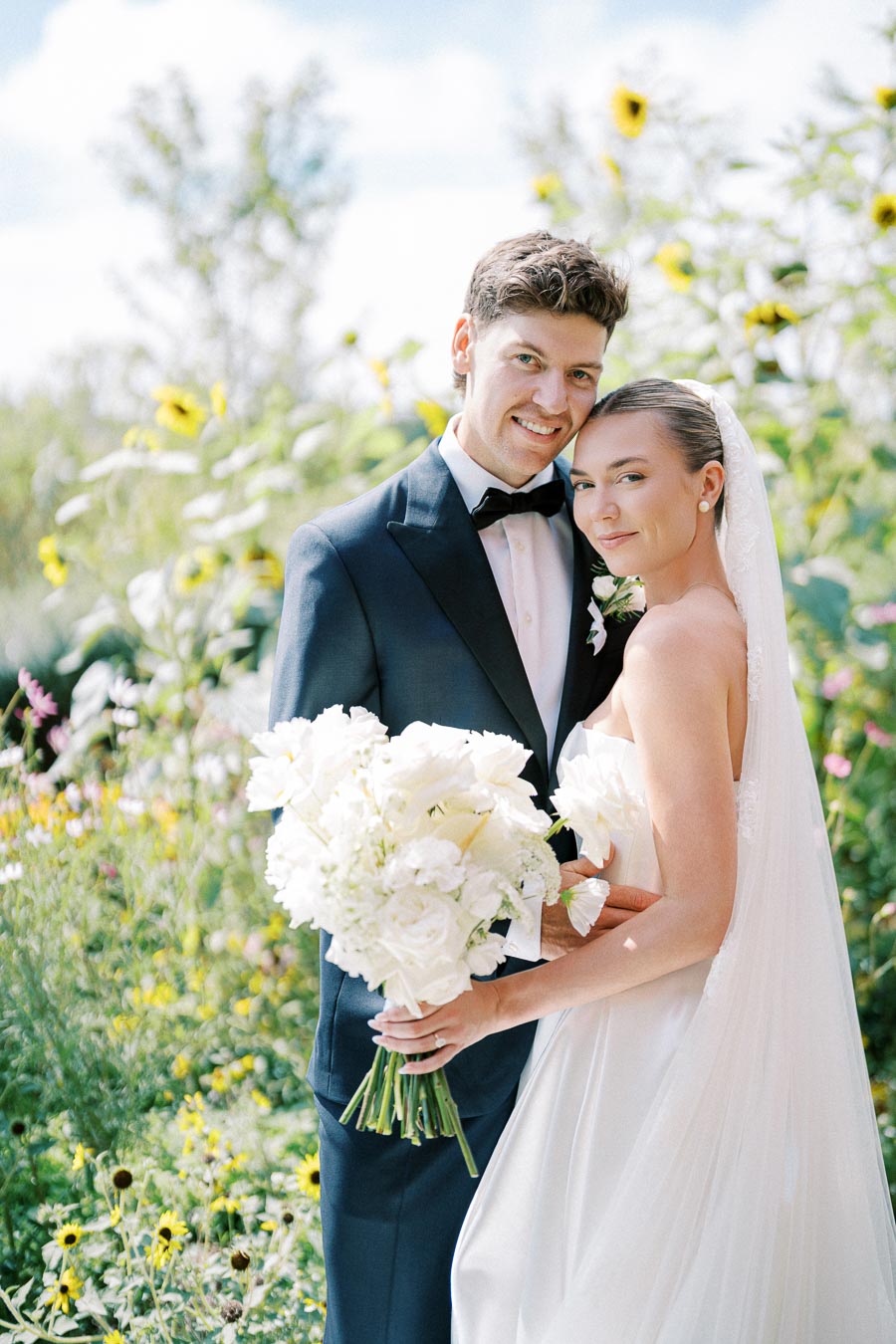 Elegant bride and groom pose together in a garden setting, holding white floral bouquets, with vibrant sunflowers in the background, capturing a perfect outdoor wedding moment.