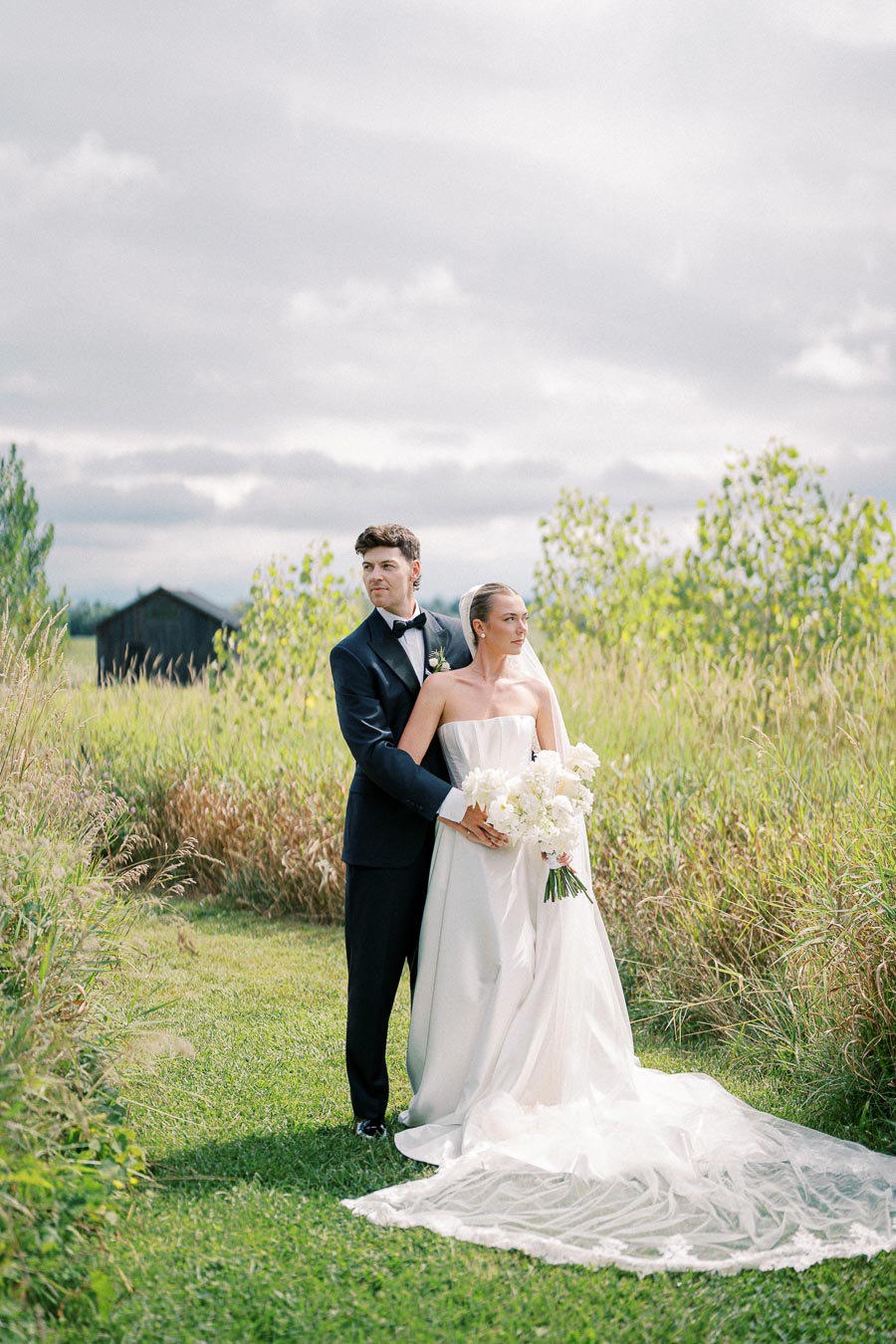 A bride and groom pose in a scenic outdoor setting, with lush greenery and cloudy skies. The bride wears a flowing white gown and holds a bouquet of white flowers, while the groom is dressed in a classic black tuxedo. Ideal for highlighting elegant, nature-inspired weddings.
