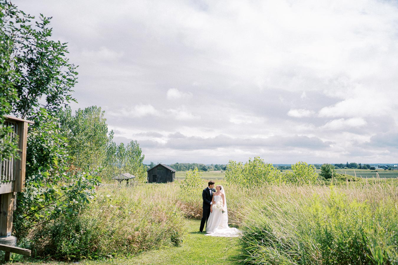 Bride and groom standing together in a lush green field with tall grasses and countryside barns in the background under a partly cloudy sky.