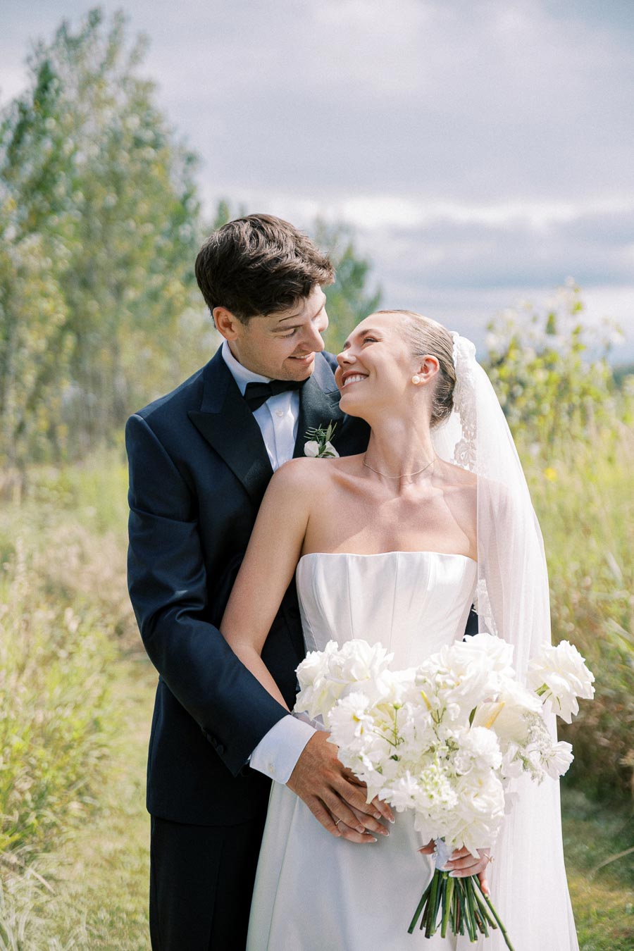 Smiling bride and groom embracing outdoors on their wedding day, with bride holding a bouquet of white flowers and greenery in the background.