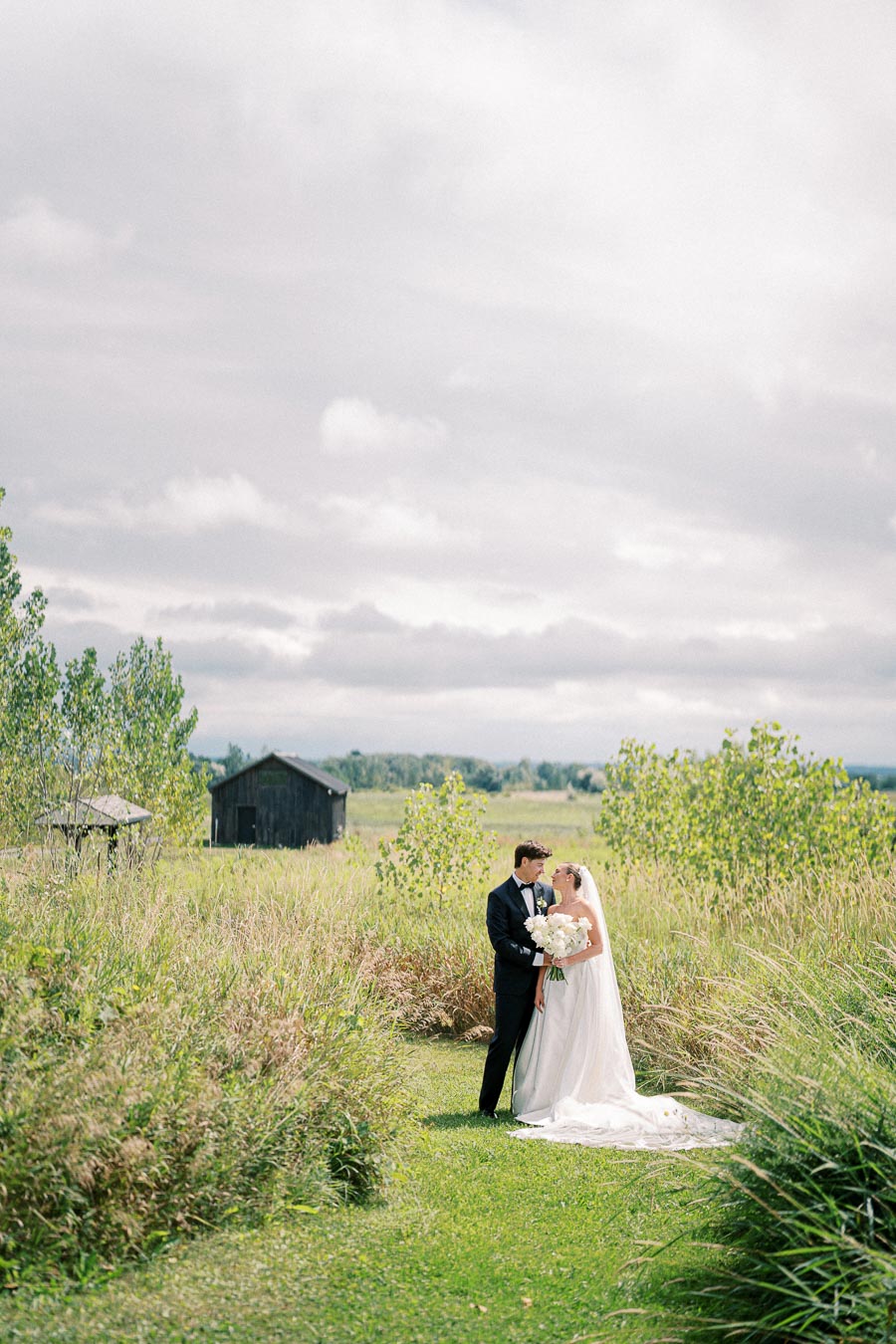 A bride and groom stand together in a picturesque meadow, surrounded by lush greenery and old barns, under a cloudy sky. The bride holds a bouquet of white flowers, both dressed elegantly for their wedding day.