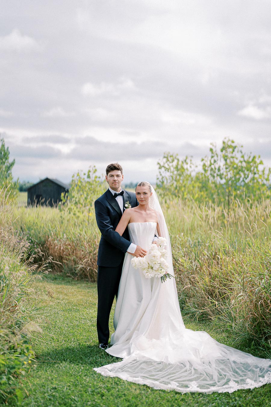 Elegant bride and groom standing in a serene outdoor setting with lush green grass and tall wild grass, under a cloudy sky, highlighting their wedding attire and peaceful countryside backdrop.
