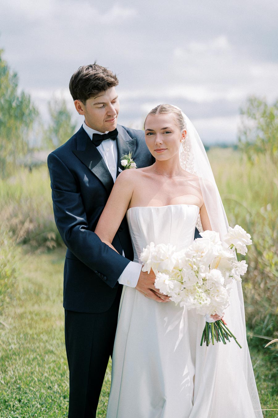 Elegant bride and groom posing outdoors in wedding attire, with the bride holding a bouquet of white flowers and the groom in a dark suit, surrounded by lush greenery and a cloudy sky backdrop.