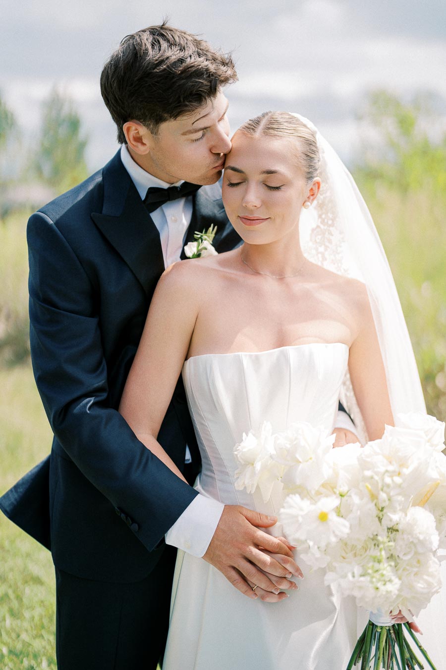 A bride and groom embrace outdoors, with the groom gently kissing the bride's forehead. The bride holds a bouquet of white flowers, and both are dressed elegantly in a wedding gown and suit.