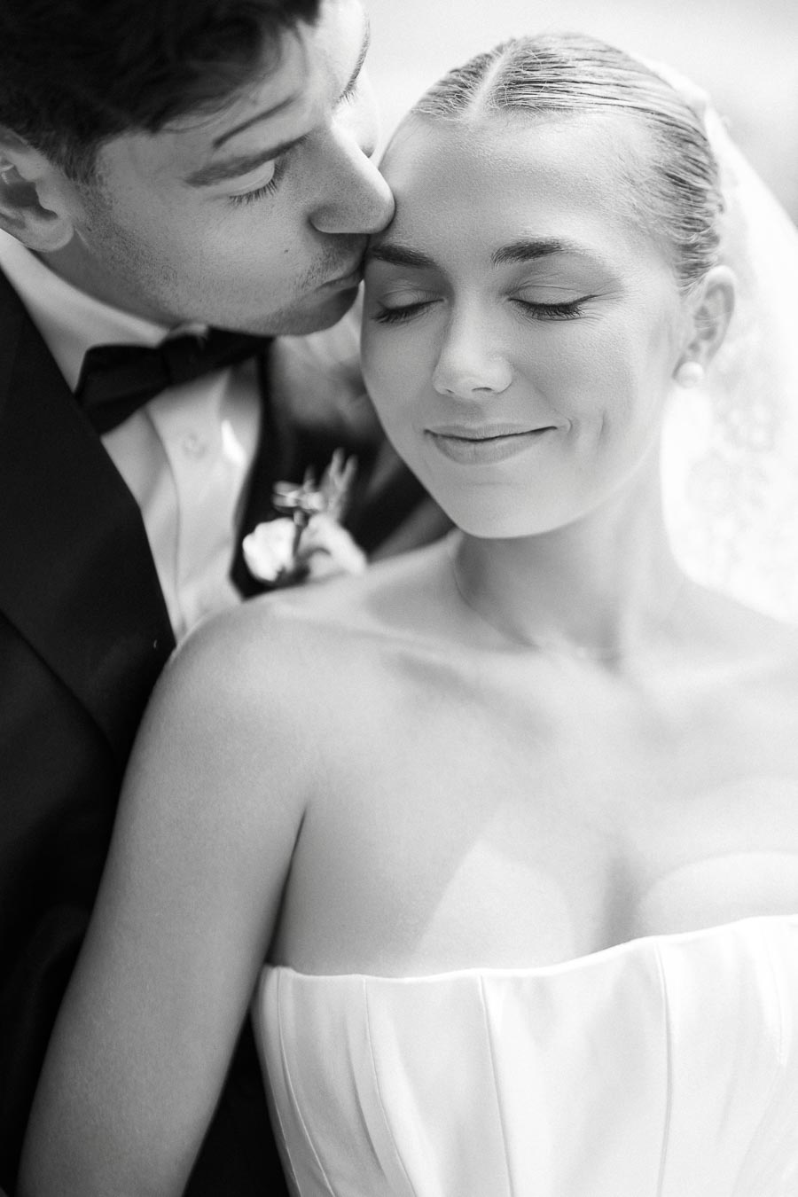 Black and white close-up of a bride in a strapless dress smiling with closed eyes, as her partner in a suit gently kisses her forehead.