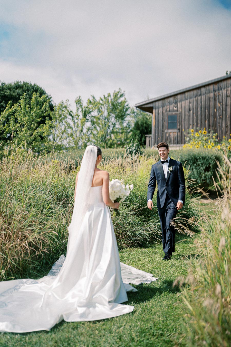 A bride in a white gown and veil holds a bouquet while walking towards a groom in a black tuxedo. They are outdoors on a grassy path surrounded by greenery, with a rustic wooden building in the background, under a partly cloudy sky.