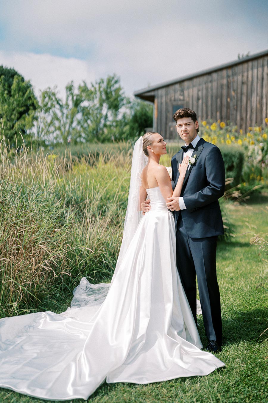 Bride and groom pose outdoors in elegant wedding attire, with the bride in a flowing white gown and veil and the groom in a dark suit. They stand on a lush green lawn with rustic wooden buildings and natural foliage in the background.