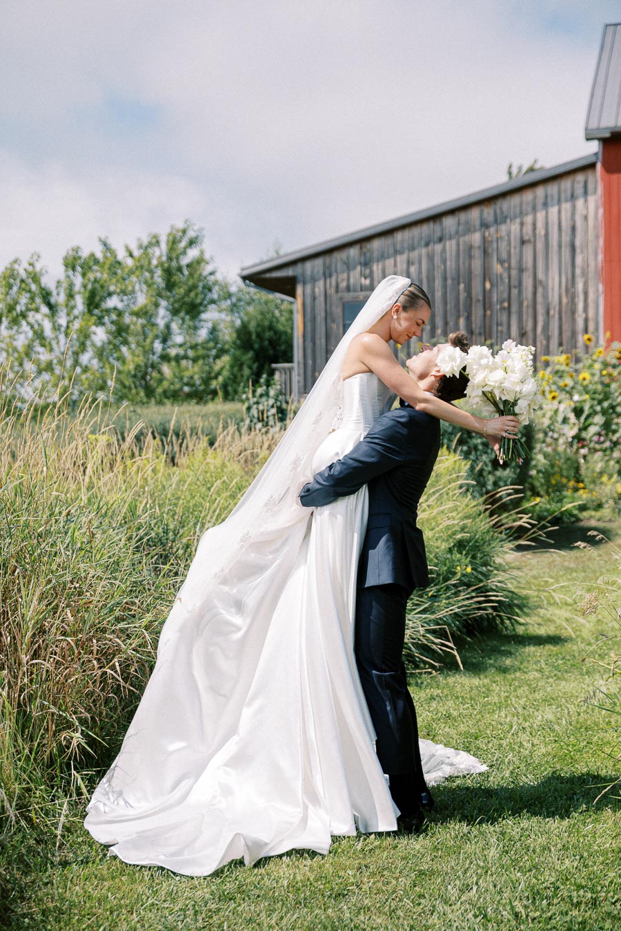 A bride and groom celebrate outdoors in front of a rustic barn, with the groom lifting the bride as she holds a bouquet of white flowers, set in a lush green garden.