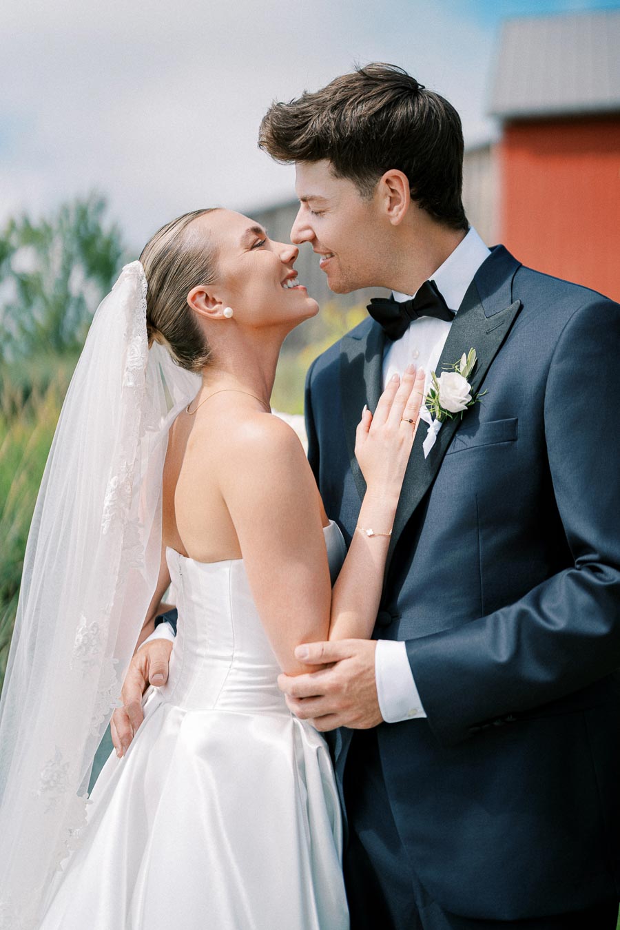 A bride and groom lovingly embrace outdoors on their wedding day, with the bride in a white gown and veil, and the groom in a dark suit with a boutonniere.
