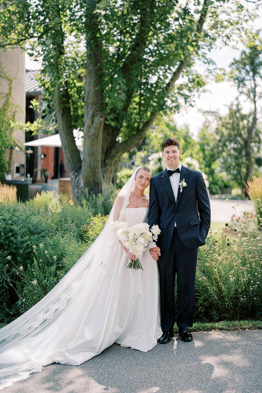 Bride and groom in elegant wedding attire posing together under a sunlit tree, surrounded by lush greenery.