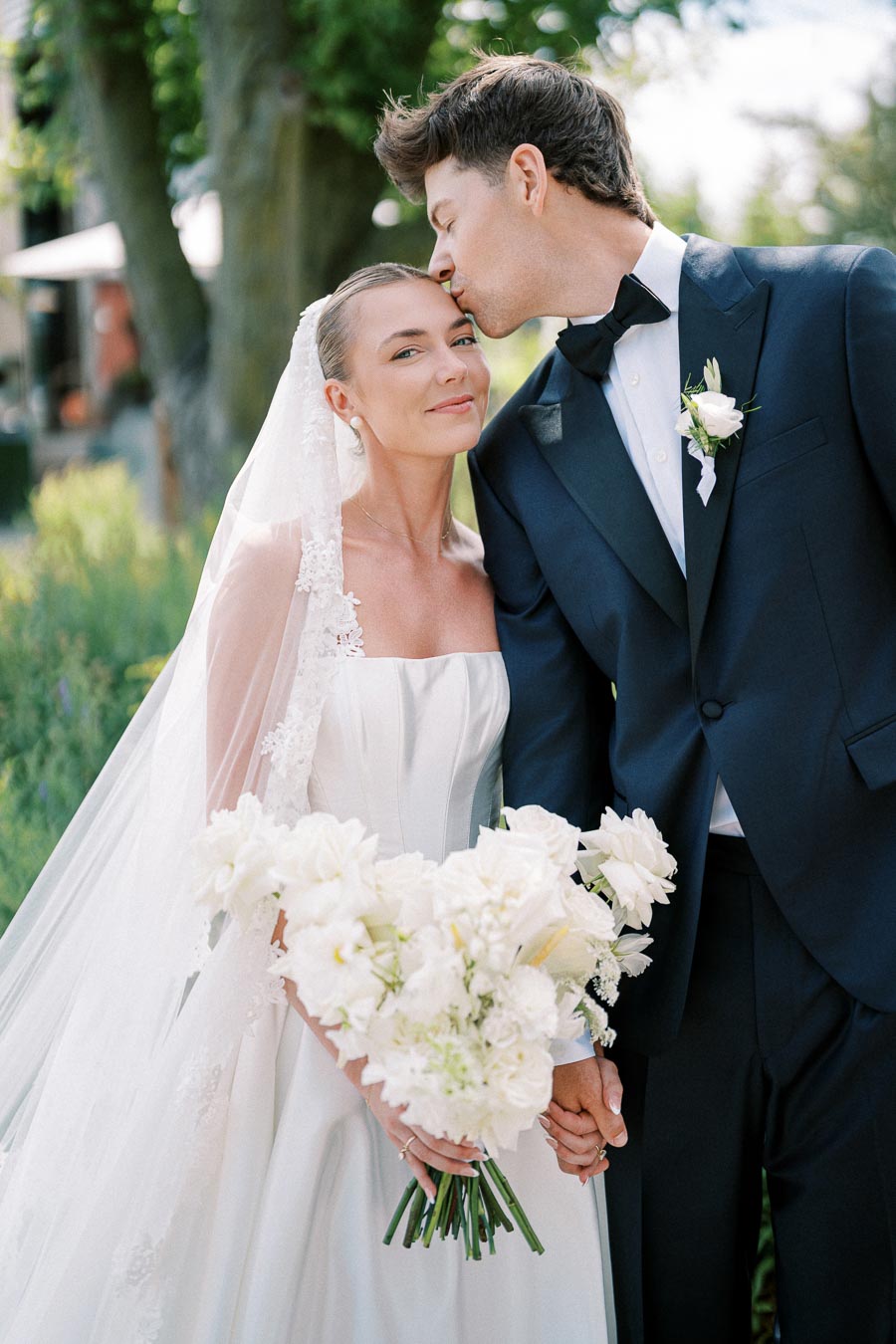 Elegant bride and groom in a loving pose, bride holding white floral bouquet, outdoor wedding setting.