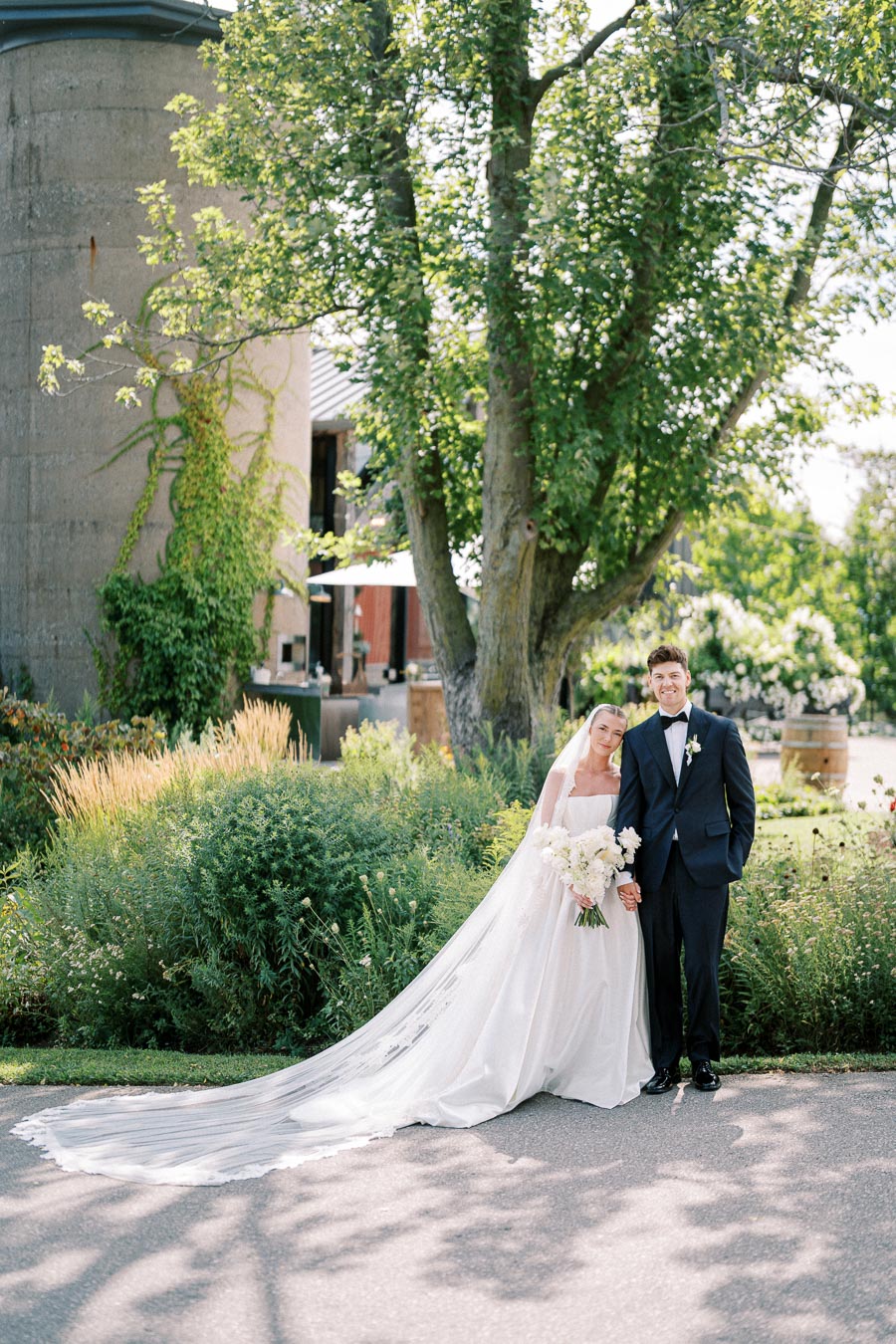 Bride and groom posing in a garden setting with lush greenery and a tree in the background, the bride wearing a flowing white gown and veil while holding a bouquet of white flowers, beside the groom in a dark suit and bow tie.