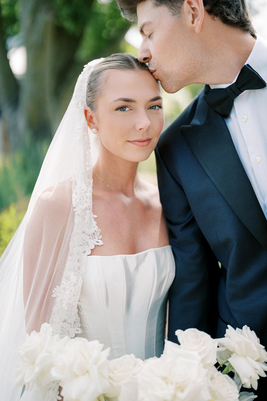 A bride in a white wedding dress and lace veil holds white roses, while standing beside a groom in a navy blue suit and black bow tie who kisses her forehead, captured in a serene outdoor setting.