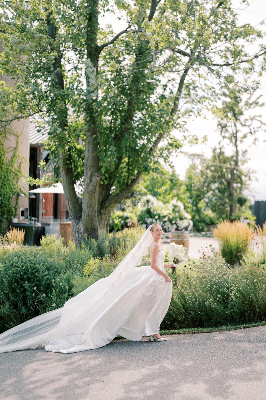 Bride wearing a flowing white wedding gown and veil, holding a bouquet, walking outdoors in a lush garden setting with trees and greenery in the background.