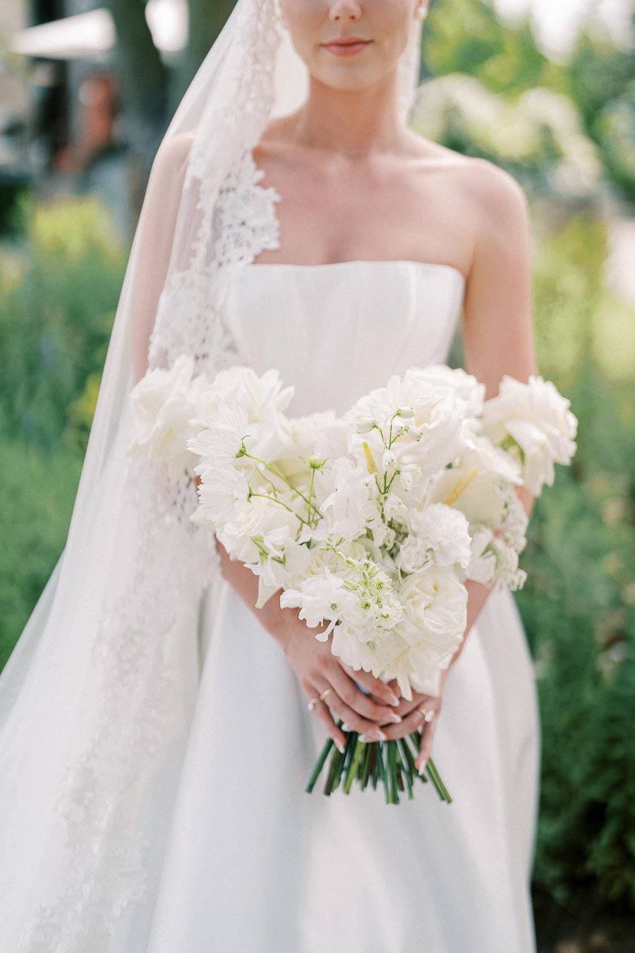 Bridal portrait featuring a woman in a strapless white wedding dress holding a bouquet of white flowers, with a lace veil flowing over her shoulders.