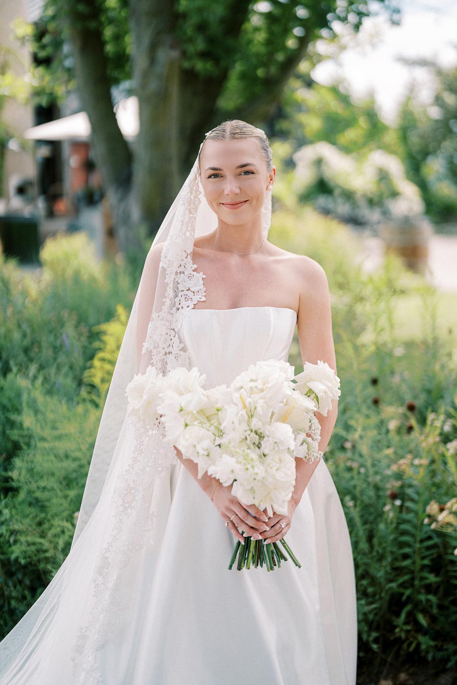 Bride in a stunning white wedding dress holding a bouquet of white flowers, standing outdoors in a lush garden setting.