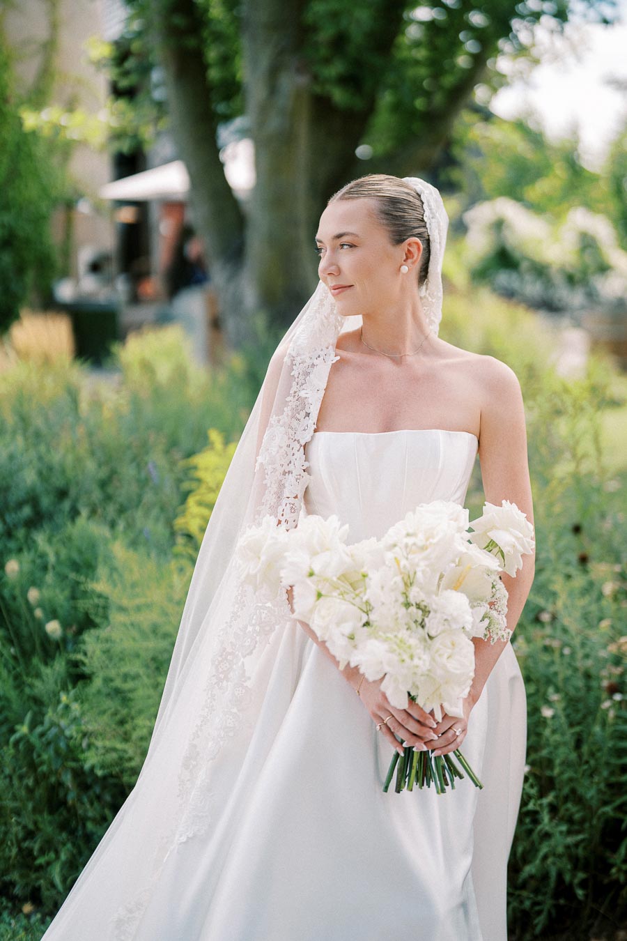 Bride in elegant white wedding gown with lace veil holding a bouquet of white flowers, standing in a lush garden setting.