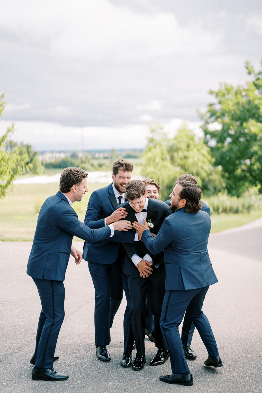 Groom surrounded by groomsmen in blue suits sharing a joyful moment outdoors on a wedding day.
