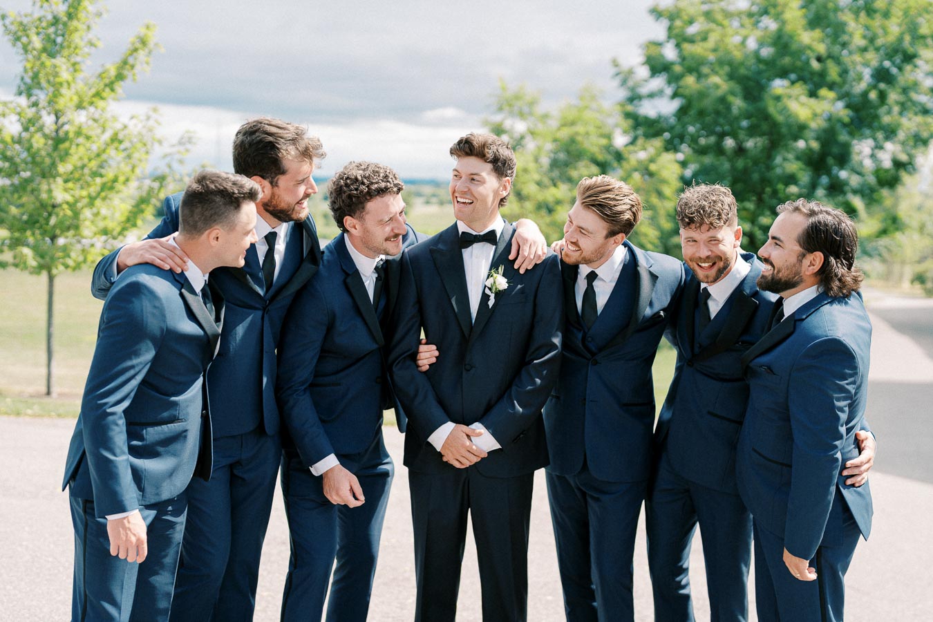 Groom and groomsmen in blue suits sharing a joyful moment outdoors, with greenery and a cloudy sky in the background.