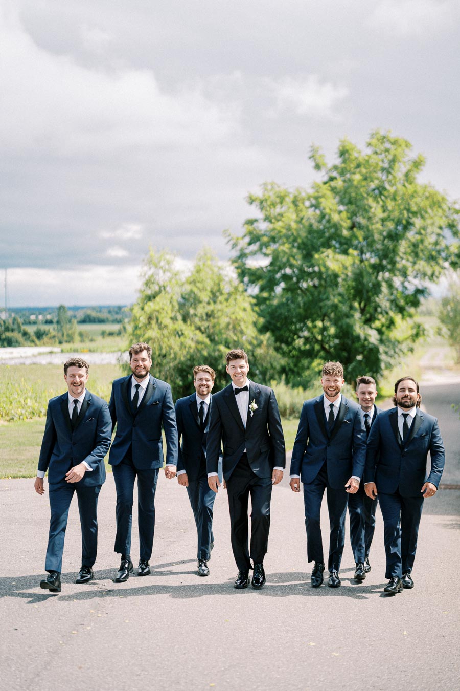 Seven groomsmen in matching suits walk confidently in an outdoor setting, with greenery and an overcast sky in the background.