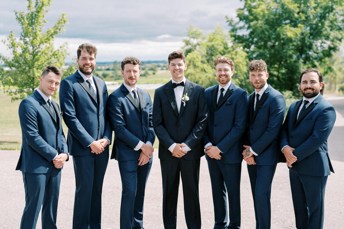 A group of seven groomsmen in matching navy blue suits stand outdoors with trees in the background, posing for a wedding photo on a sunny day.