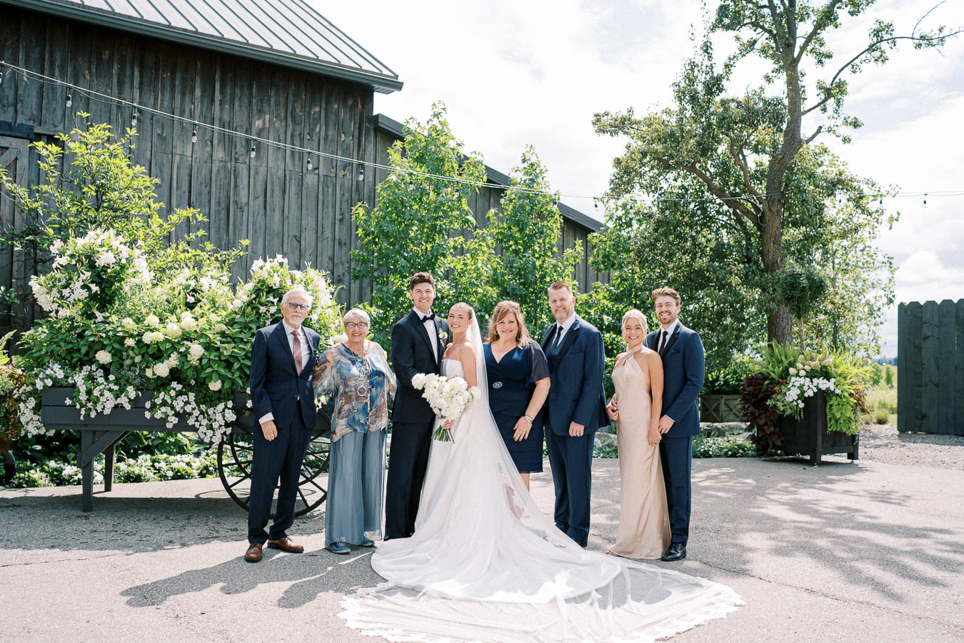 A joyful wedding group photo, featuring a bride in a white gown and groom in a black suit surrounded by family members, outside a rustic venue adorned with flowers and greenery.