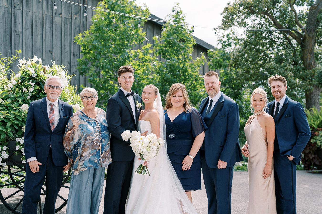 A wedding group photo featuring a joyful bride and groom surrounded by family members, all dressed formally, standing outdoors with lush greenery and floral arrangements in the background.