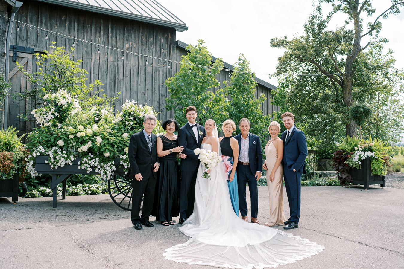 A happy wedding group poses in formal attire outside a rustic barn with lush greenery and floral arrangements. The bride in a white gown holds a bouquet, surrounded by smiling family members and friends in elegant dresses and suits.