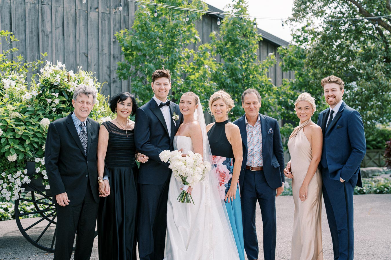 A group of people dressed in formal attire posing for a wedding photo outdoors, with lush greenery and flowers in the background.