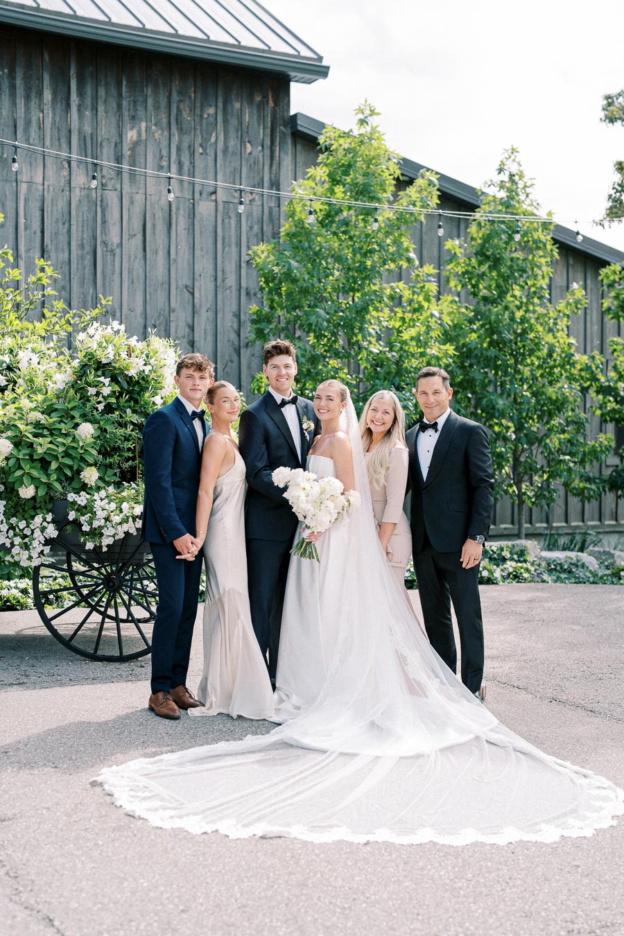 A bride and groom surrounded by friends and family, all dressed elegantly, pose in front of a rustic wooden building adorned with greenery and flowers on a sunny day.