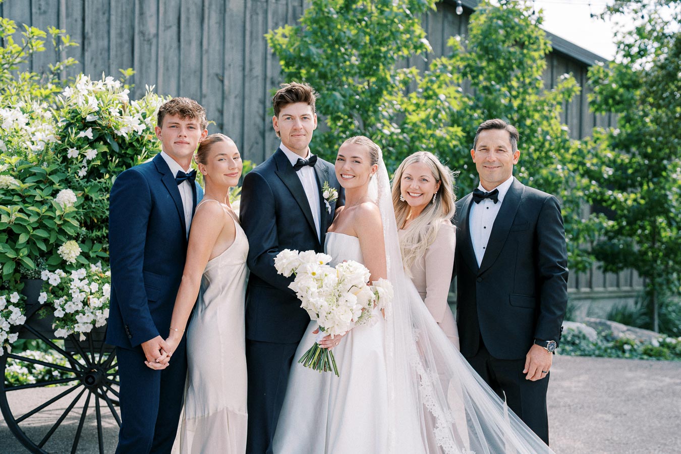 Wedding party posing outdoors, with bride in white gown holding bouquet, groom in suit, surrounded by four elegantly dressed people, lush greenery and rustic background.