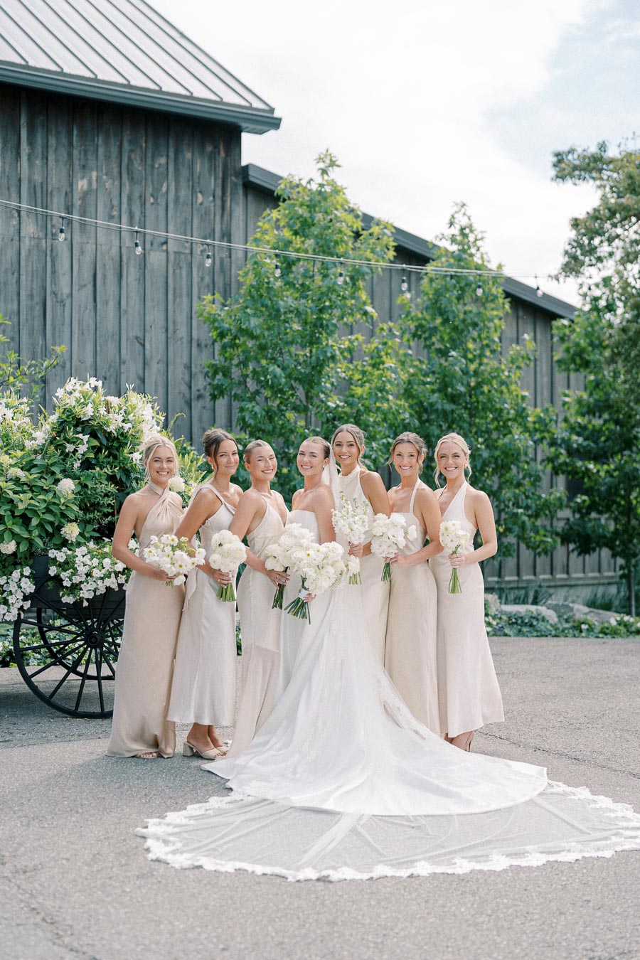 Bridal party posing outdoors with bouquets in front of rustic wooden building, featuring bride in white gown and bridesmaids in neutral dresses.