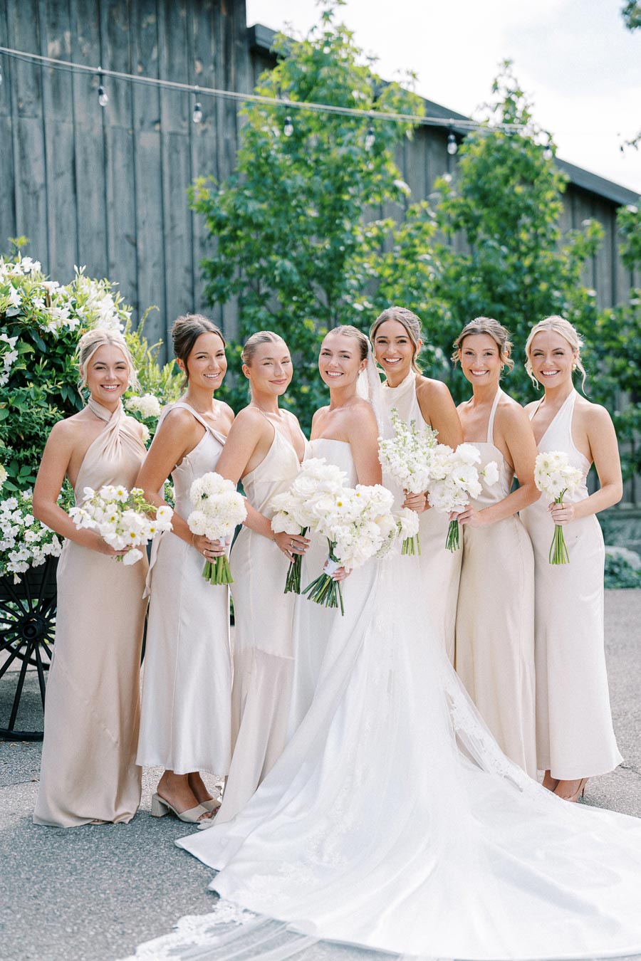 Seven bridesmaids in elegant cream dresses holding white floral bouquets, pose together outdoors in front of a rustic wooden building, highlighting a beautiful wedding scene.