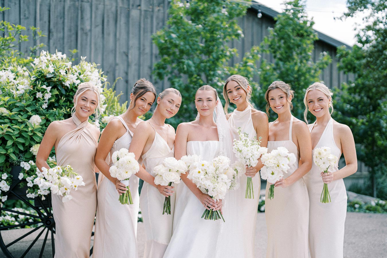 Bridal party posing outdoors in pastel dresses, each holding white floral bouquets, with greenery backdrop and rustic wooden wall.