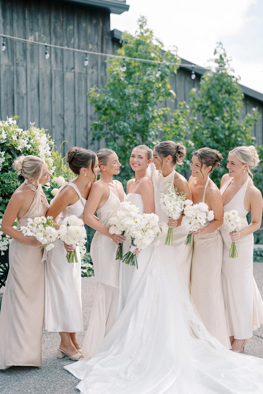 Bride and bridesmaids in elegant white dresses holding bouquets of white flowers, posing happily together outdoors, perfect for a summer wedding.