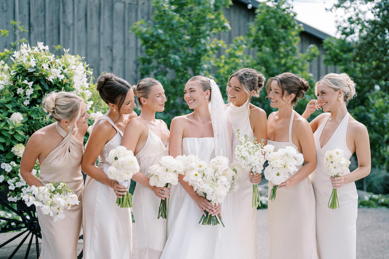 Bridal party posing outdoors with the bride and bridesmaids in elegant pastel dresses, each holding a bouquet of white flowers, surrounded by lush greenery.