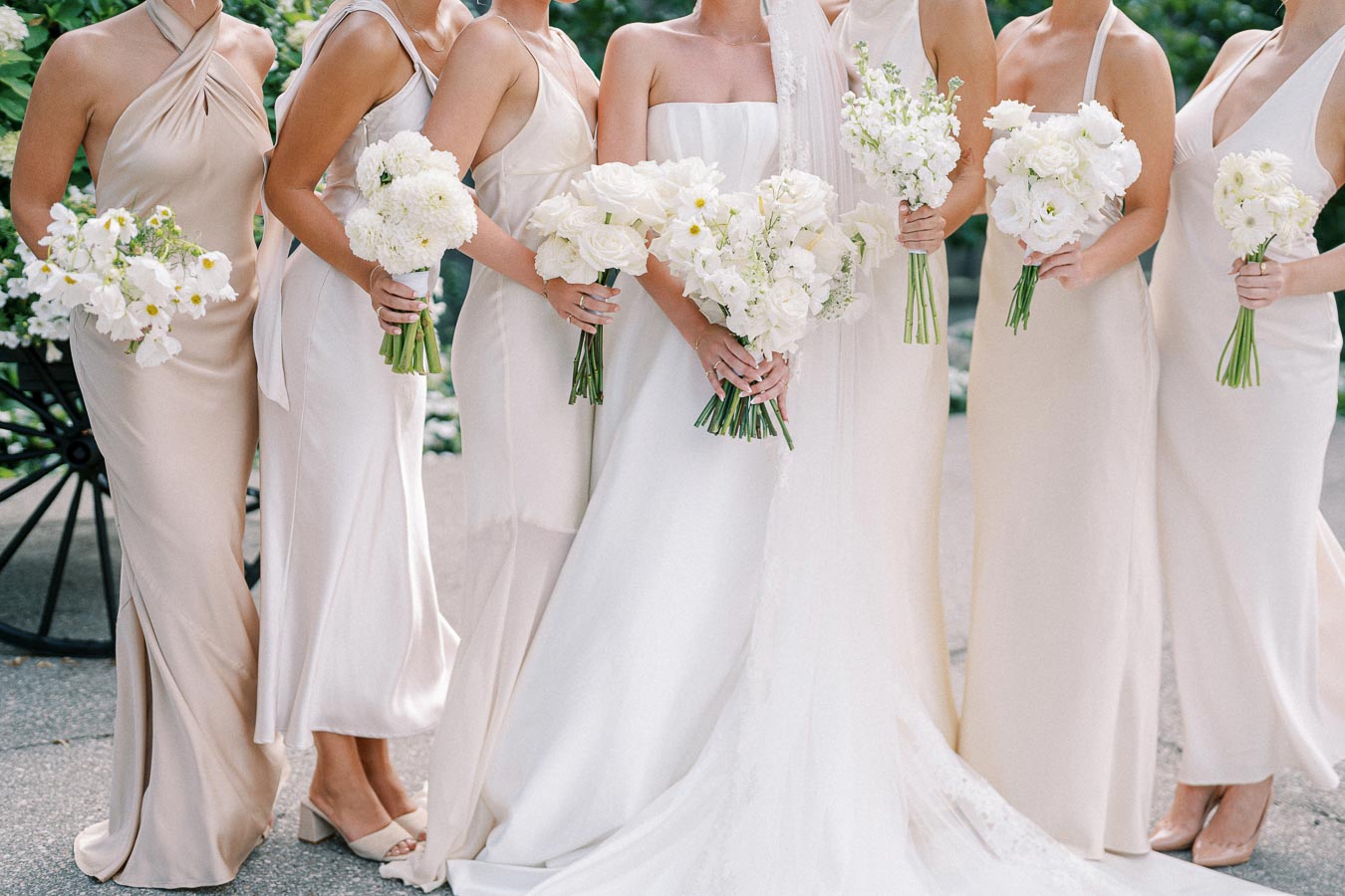 Bridal party with elegant cream dresses holding white floral bouquets, standing outdoors on a sunny day.