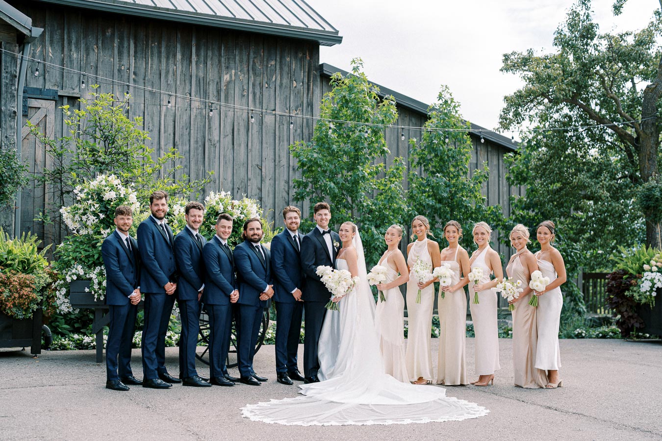 Bridal party posing outdoors in front of rustic barn, featuring groom and groomsmen in navy suits and bride with bridesmaids in light dresses, all holding white floral bouquets.
