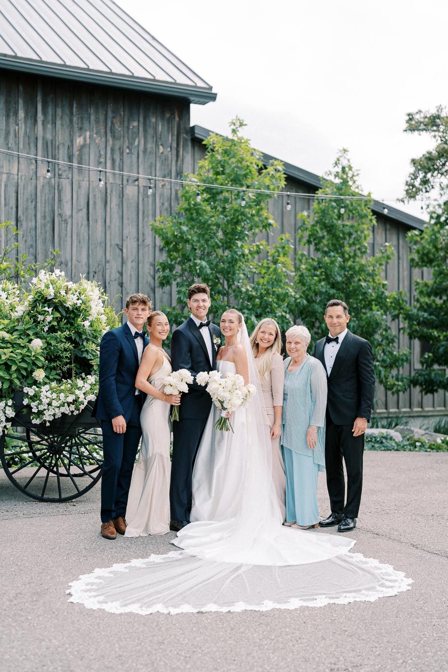 Wedding party standing in front of a rustic barn, featuring a bride in a flowing white gown and veil holding a bouquet, surrounded by family members dressed in formal attire, with decorative greenery and white flowers in the background.