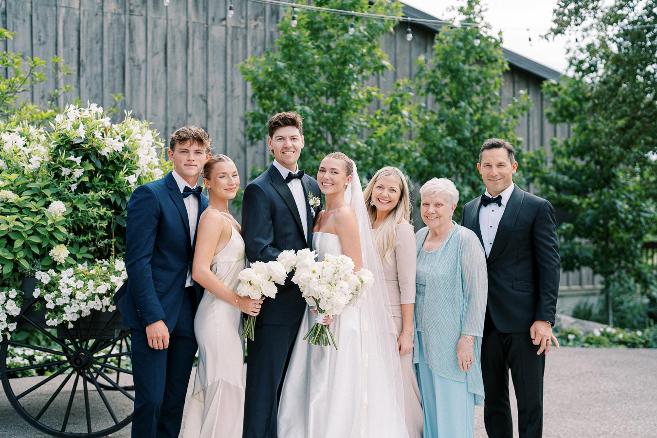 Wedding party in elegant attire posing outdoors with bouquets of white flowers, set against a rustic wooden barn and lush greenery.