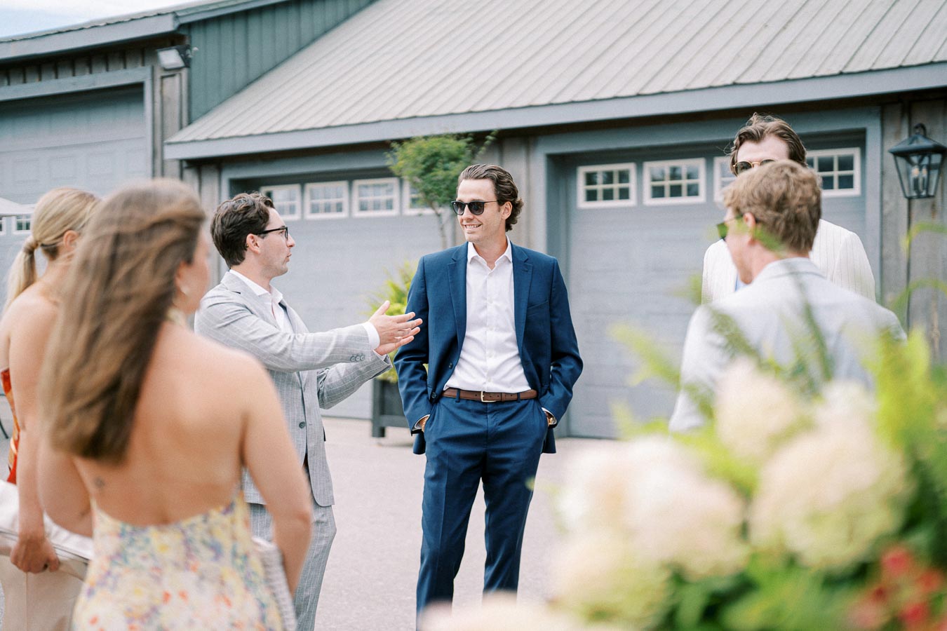 Group of people in formal attire having a conversation outdoors in front of a modern building with garage doors, highlighting an elegant event or social gathering.