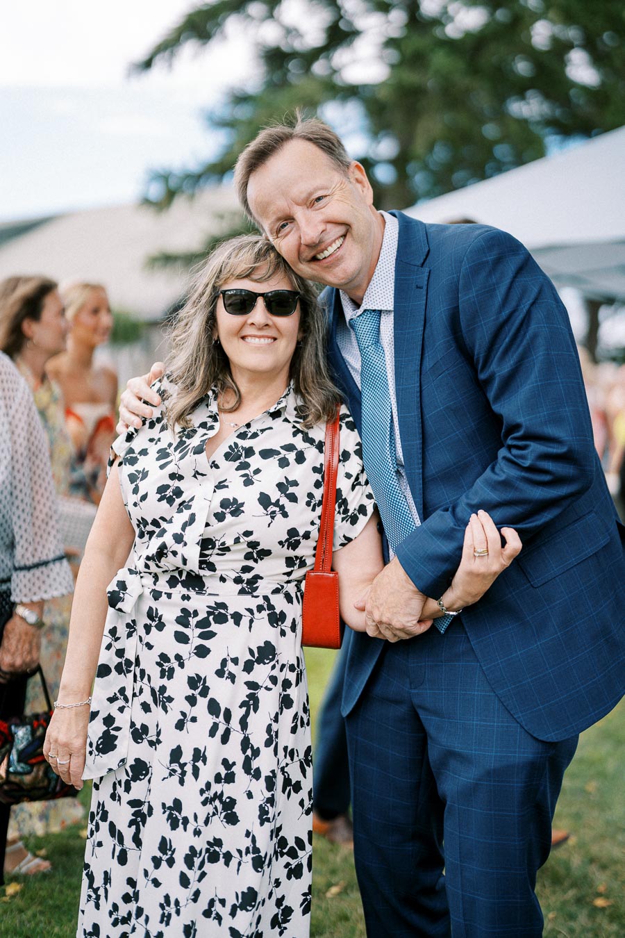 Smiling couple pose together outdoors at a social event; woman in a floral dress and sunglasses, man in a blue suit with a patterned tie.