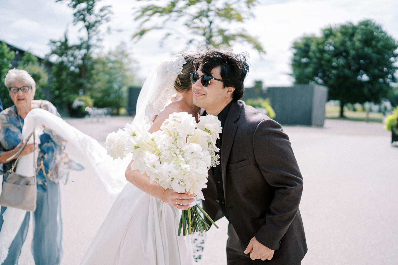 Bride wearing a white dress and holding a bouquet of white flowers is embracing a man in a dark suit and sunglasses outdoors, with trees and a sunny sky in the background.