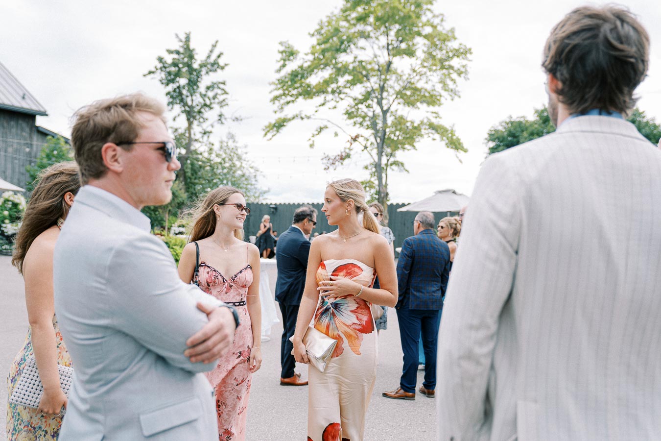 A group of elegantly dressed people socializing outdoors at a daytime event, with lush greenery and trees in the background, under a cloudy sky.