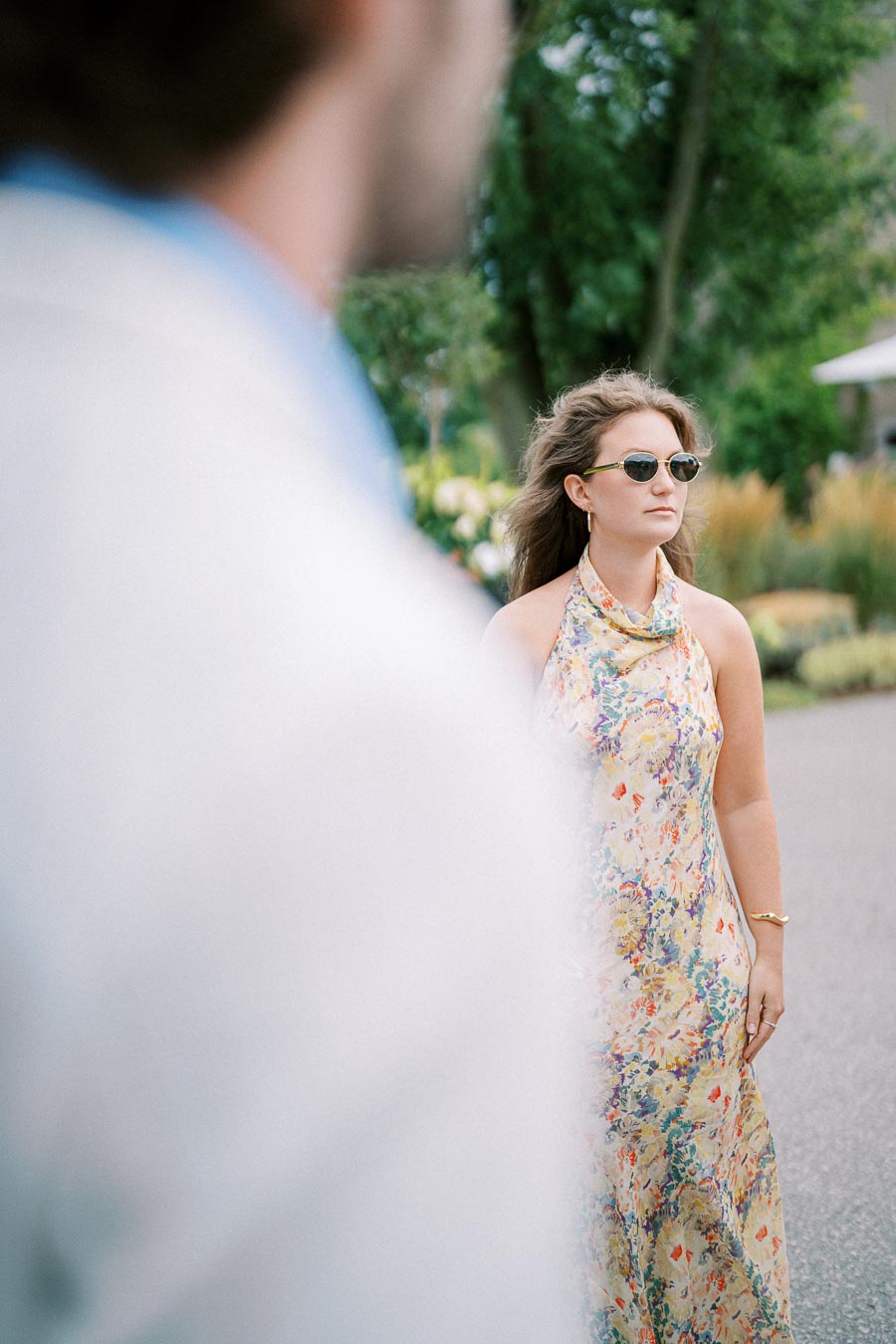 A woman in a floral dress and sunglasses stands outdoors, looking into the distance on a sunny day, with a blurred figure in the foreground.