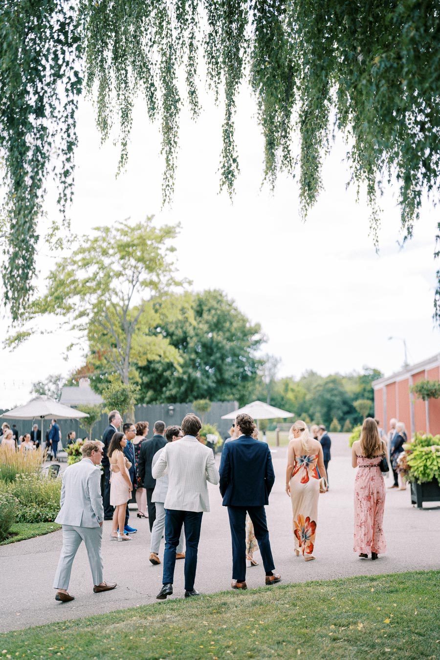 A group of elegantly dressed people gathered outdoors at a garden wedding reception under a canopy of lush green trees.