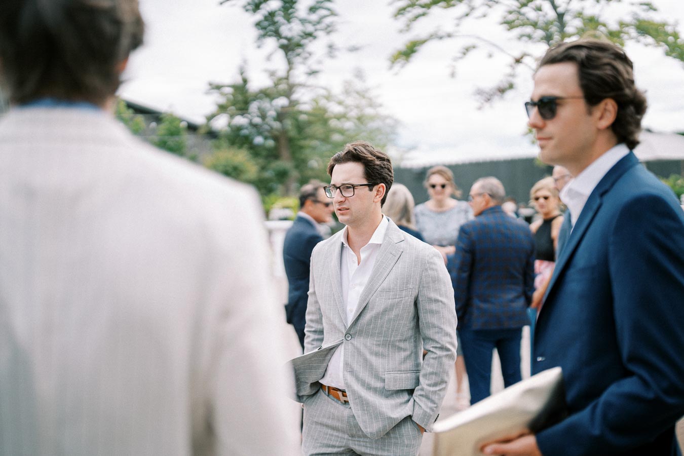 A group of people in formal attire conversing outdoors at an event; a man in a light gray suit stands in focus, surrounded by other attendees in suits and sunglasses, with trees and sky in the background.